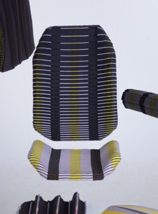 Eine Sitzfläche und Rückenlehne aus gewebtem Textil mit schwarzen, gelben, weißen und grauen Streifen, die vor einem schlichten weißen Hintergrund aufgehängt sind.
