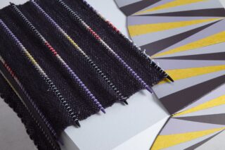 Eine Nahaufnahme eines schwarzen strukturierten Stoffes mit bunten Perlen auf Stricknadeln, neben einem geometrischen Muster in Gelb-, Grau- und Lilatönen.