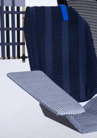 Ein moderner Stuhl mit blau-schwarz gestreifter Rückenlehne und einer flachen, hellgrauen Sitzfläche mit linearem Muster vor einem minimalistischen Hintergrund mit geometrischen Elementen.