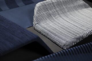 Nahaufnahme mehrerer gefalteter Stoffe in unterschiedlichen Blau- und Grautönen, die unterschiedliche gewebte Texturen und Muster auf dunklem Hintergrund zeigen.