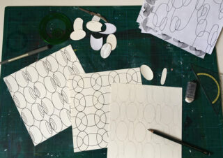 Ein Arbeitsbereich mit gemustertem Papier, Zeichenwerkzeugen, Papierausschnitten und einem Bleistift auf einer grünen Schneidematte.