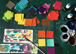Stapel bunter Papierquadrate, Flaschen mit schwarzer Farbe oder Tinte, ein Pinsel, Wasserfarben und eine Palette sind auf einer grünen Schneidematte angeordnet.
