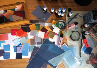 Ein Arbeitsbereich mit farbigen Papierquadraten, Markern, Tintenfässern, einem Pinsel, einem Glasgefäß und verstreuten Kunstutensilien auf einem Holztisch.