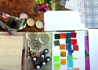 Künstlerbedarf auf einem Holztisch, darunter Pinsel, Farben, Gläser, gemustertes Papier, farbige Farbmuster, ein Tuch und eine rosa Blume in einer Vase.