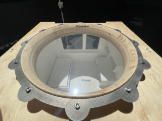 Ein rundes Glasfenster mit Holzrahmen spiegelt einen weißen Innenraum mit Regalen und einem Fenster wider, montiert auf einer hellen Holzoberfläche mit der Aufschrift „EVANS“.