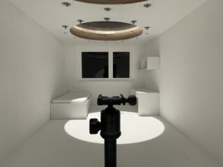 Ein Kamerastativ steht in der Mitte eines minimalistischen weißen Raums mit einem runden Oberlicht, eingebauten Betten, Regalen und einem Fenster am anderen Ende.