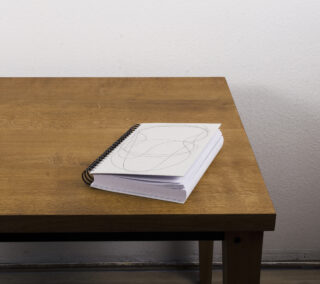 Ein spiralgebundenes Notizbuch mit abstrakten schwarzen Strichzeichnungen auf dem Einband liegt auf einem Holztisch vor einer schlichten weißen Wand.