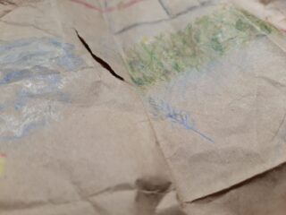 Eine Nahaufnahme eines zerrissenen Stücks braunem Papier mit schwachen Buntstiftzeichnungen, darunter grünes Gras und blaue Markierungen.