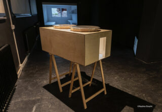 Eine rechteckige Holzbox mit zwei runden, mit Maschen bedeckten Öffnungen steht auf einem Holzständer in einem schwach beleuchteten Raum mit projizierten Bildern an den Wänden.