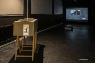 In einem dunklen Raum steht ein Holzgerät auf einem Ständer. An der gegenüberliegenden Wand werden digitale Bilder projiziert. Auf dem Boden steht „©Nadine Bracht“.