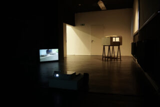 Ein schwach beleuchteter Raum mit einem Videoprojektor auf dem Boden, einer Leinwand, auf der ein Bild angezeigt wird, und einem kleinen Architekturmodell auf einem Ständer neben einer weißen Tür.