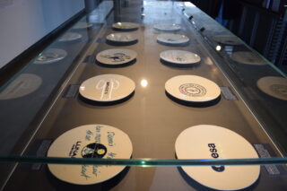 Glasvitrine mit einer Sammlung von Gedenktafeln, jede mit einem eigenen Logo und Design, in Reihen auf einer flachen Oberfläche angeordnet.