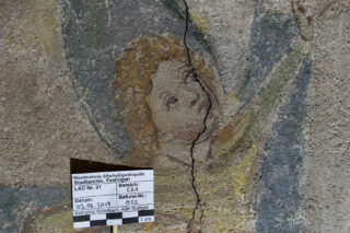 Ein rissiges Wandgemälde einer Engelsfigur, unter dem Kunstwerk befindet sich eine Referenzkarte zur Größenbestimmung und Identifizierung.