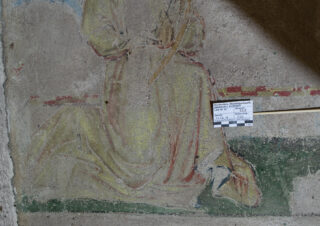 Verblasstes Wandgemälde, das eine kniende, in eine Robe gehüllte Gestalt mit gefalteten Händen zeigt, teilweise beschädigt, neben einer Maß- und Datenkarte als Referenz.