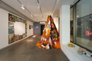 Eine Kunstgalerie zeigt verschiedene Gemälde an weißen Wänden und in der Mitte eine große, farbenfrohe, stoffbespannte Kegelskulptur, während auf einem Fensterbrett kleine Skulpturen stehen.