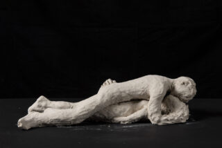 Zwei Tonfiguren mit rauer Textur sind auf einer flachen Oberfläche positioniert, wobei vor einem schwarzen Hintergrund eine Figur auf der anderen liegt.