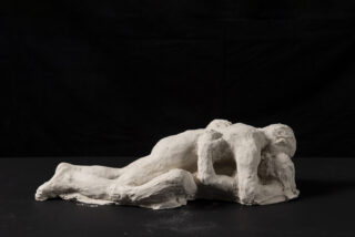 Eine Skulptur aus weißem Ton, die zwei liegende, sich umarmende menschliche Figuren vor einem dunklen Hintergrund zeigt.