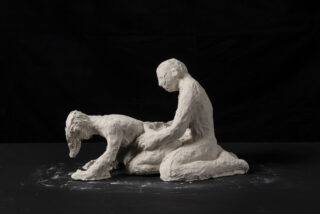 Eine grob strukturierte weiße Skulptur zweier menschlicher Figuren; eine kniet aufrecht, während die andere auf allen Vieren steht, beide vor einem schwarzen Hintergrund.