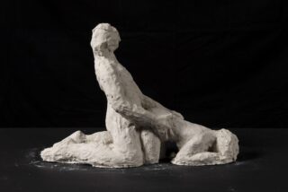 Eine grobe Tonskulptur zeigt vor einem schwarzen Hintergrund zwei gesichtslose menschliche Figuren in einer sexuellen Stellung.