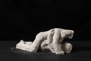 Eine Skulptur aus weißem Ton stellt zwei übereinander angeordnete menschliche Figuren auf dunklem Hintergrund dar.
