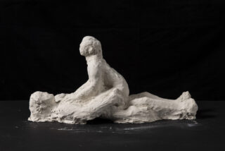 Eine grobe Ton-Skulptur zweier menschlicher Figuren, die innig miteinander interagieren, wobei eine Figur auf der anderen sitzt, vor einem schwarzen Hintergrund.