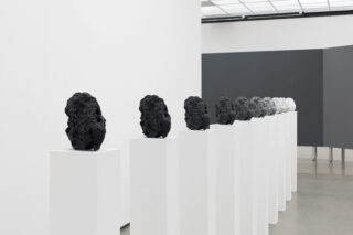 Neun strukturierte Skulpturen in verschiedenen Farbtönen von Schwarz bis Weiß werden in einer Reihe auf weißen Sockeln in einem minimalistischen Galerieraum ausgestellt.