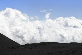 Felsiges, dunkles Vulkangelände im Vordergrund mit dicken, weißen Wolken im Hintergrund und einem klaren blauen Himmel darüber.