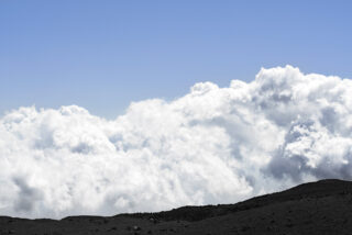 Ein blauer Himmel mit großen, weißen Wolken über einem dunklen, felsigen Gelände im Vordergrund.