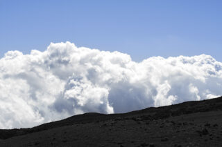Ein dunkler, felsiger Vordergrund kontrastiert mit großen, bauschigen weißen Wolken und einem klaren blauen Himmel im Hintergrund.