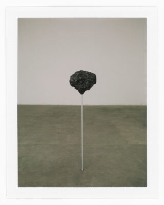 Ein schwarzer Stein schwebt über dem Boden, gestützt von einer dünnen vertikalen Stange in einem ansonsten leeren Raum mit Betonboden und einer hellen Wand.