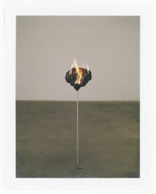 Ein dunkler, unregelmäßig geformter Stein hängt an einer dünnen vertikalen Stange und brennt mit sichtbaren Flammen vor einem schlichten, neutralen Hintergrund.