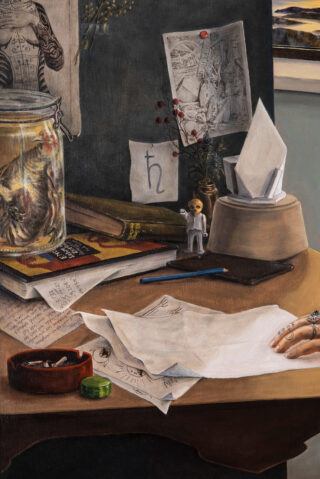 Eine Hand ruht auf Papieren auf einem überfüllten Schreibtisch mit Skizzen, Büchern, einer Spielfigur, einem Glas, Bleistiften und einem Taschentuchhalter; im Hintergrund sind angeheftete Zeichnungen und ein Fenster zu sehen.