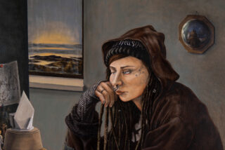 Eine Person mit Dreadlocks, Gesichtstattoos und Piercings, die einen braunen Kapuzenpullover trägt, sitzt im Haus neben einem Fenster mit Blick auf den Sonnenuntergang und einem runden Spiegel, in dem sich ihr Profil spiegelt.