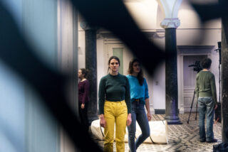 Vier Personen stehen in einem Raum mit Marmorboden und Säulen; zwei Frauen blicken nach vorne, eine Frau steht seitlich, und im Hintergrund bedient ein Mann eine Kamera. Ein schwarzer Gegenstand verdeckt teilweise die Sicht.