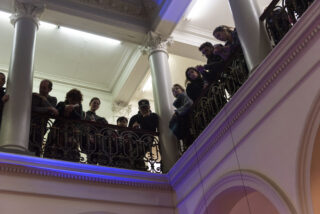 Eine Gruppe von Menschen steht auf einem Balkon in einem Gebäude mit verzierten Säulen und blickt über ein dekoratives Geländer nach unten.