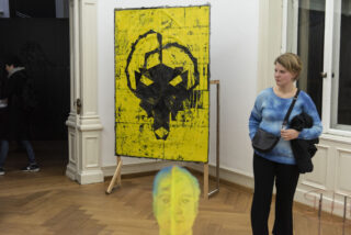 Eine Frau steht in einer Kunstgalerie neben einem gelben abstrakten Gemälde mit schwarzen geometrischen Formen. Im Vordergrund ist ein weiteres Kunstwerk mit einem Gesicht zu sehen.