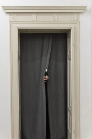 Eine Hand mit einer Bierflasche kommt hinter einem dunkelgrauen Vorhang hervor, der in einem verzierten Türrahmen hängt.