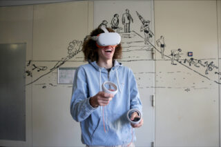 Eine Person, die ein VR-Headset trägt und Controller in der Hand hält, lächelt vor einer Wand mit einem Wandgemälde zur menschlichen Evolution und zum wissenschaftlichen Fortschritt.