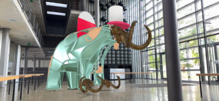 In einem modernen Gebäude mit Glaswänden und Elementen im Industriedesign ist eine digitale Kunstinstallation eines stilisierten Wollmammuts mit rosa Hut ausgestellt.