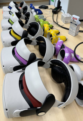 Eine Reihe von Virtual-Reality-Headsets mit verschiedenfarbigen Frontplatten und passenden Controllern sind auf einem Tisch in der Nähe einiger elektronischer Geräte und einer Saftpackung angeordnet.