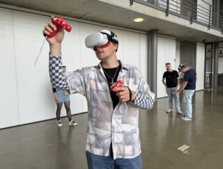 Eine Person mit einem VR-Headset und Controllern steht im Innenbereich, im Hintergrund sind drei weitere Personen.