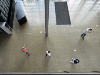 Von oben betrachtet stehen vier Personen mit etwas Abstand voneinander auf einem polierten Betonboden in einem großen Innenraum.