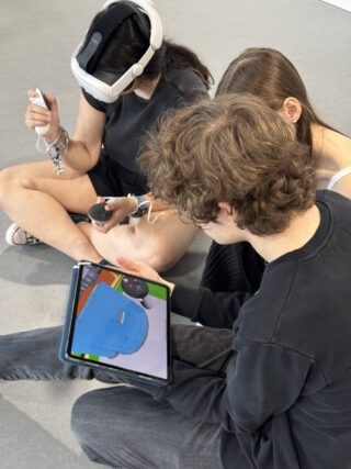 Drei Personen sitzen auf dem Boden; eine trägt ein VR-Headset, während eine andere ein Tablet hält, auf dem ein 3D-Modell angezeigt wird.