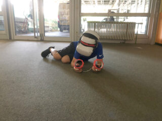 Eine Person kniet auf einem Teppichboden, trägt ein VR-Headset und hält zwei Handsteuerungen und scheint mit einer virtuellen Umgebung zu interagieren.