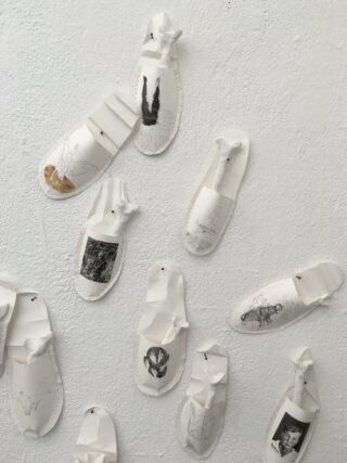 An eine strukturierte weiße Wand sind weiße Papierskulpturen gepinnt, die wie Schuhe aussehen. Jede Skulptur enthält unterschiedliche Skizzen oder Bilder.