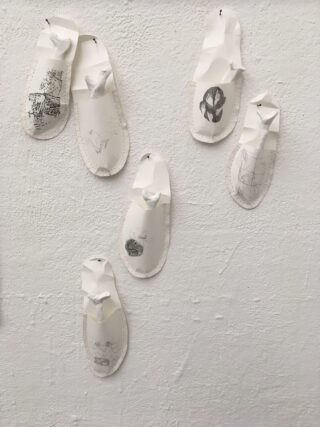 An einer weißen strukturierten Wand sind sechs Papierausschnitte mit Bleistiftzeichnungen befestigt. Jeder hat die Form einer Sandale mit einem erhabenen Abschnitt, der einen Flip-Flop-Riemen darstellt.