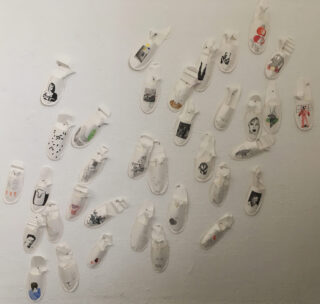 Mehrere weiße Papierschuhausschnitte mit unterschiedlichen handgezeichneten Motiven sind verstreut auf einer weißen Wand angebracht.