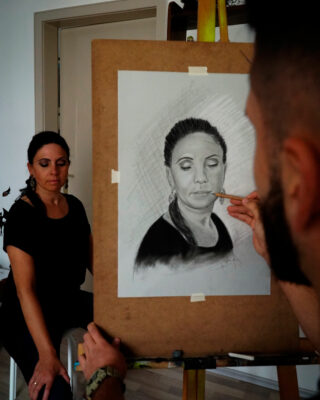 Eine Frau posiert mit geschlossenen Augen, während ein Künstler in einem gut beleuchteten Raum ihr Porträt auf einer Staffelei skizziert.