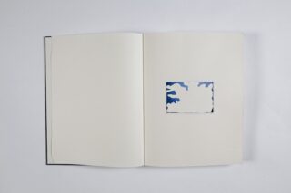 Ein offenes Buch mit leeren Seiten; die rechte Seite weist einen kleinen rechteckigen Ausschnitt auf, unter dem sich ein blau-weißes Wolkenmuster befindet.