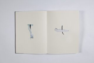 Ein offenes Buch mit zwei leeren Seiten, jede mit einer ausgeschnittenen Form; der linke Ausschnitt ist rechteckig, der rechte Ausschnitt ist unregelmäßig und darunter ist teilweise ein spiralförmiger Gegenstand sichtbar.